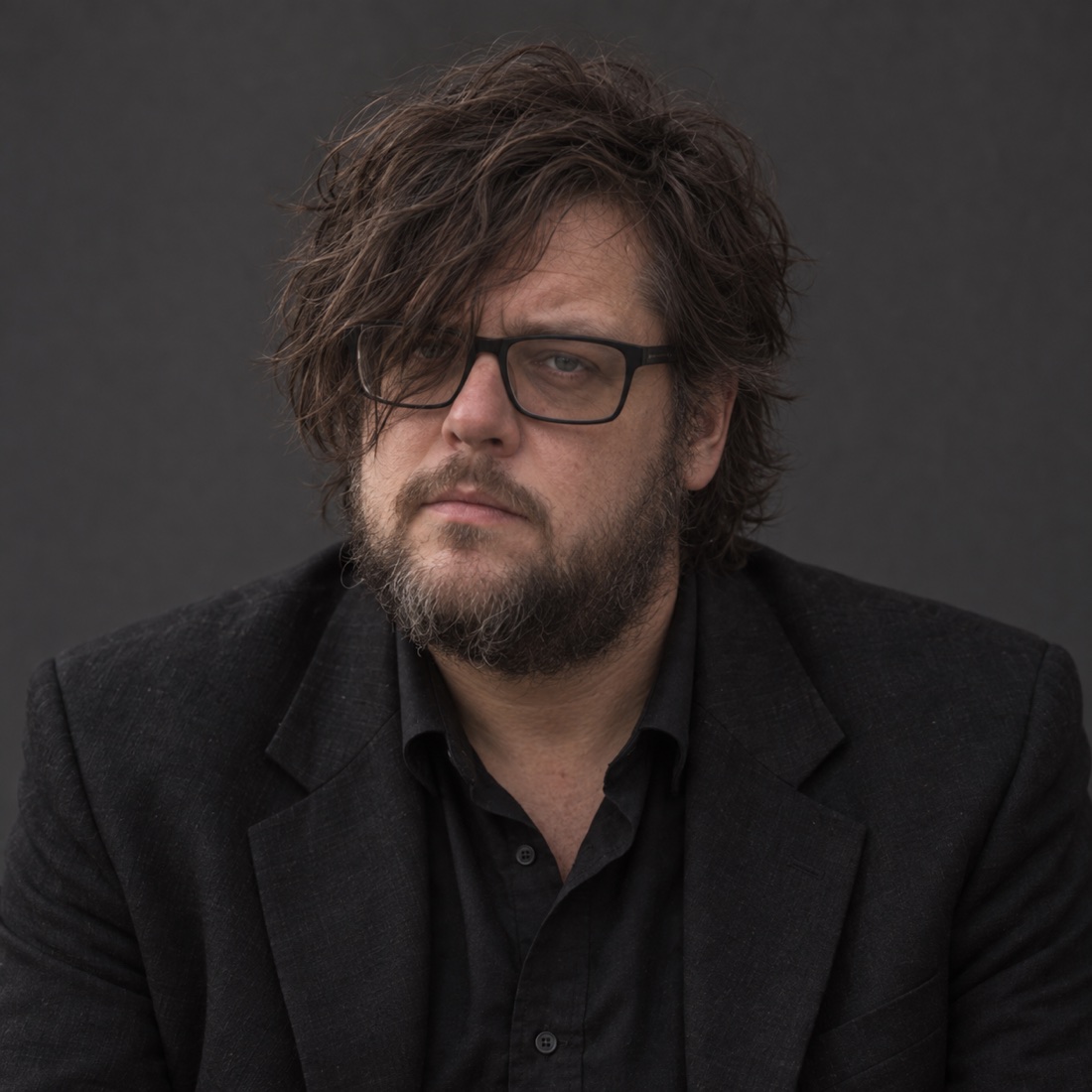 Chain Wallace with tousled dark hair, black glasses, a short beard and a black jacket